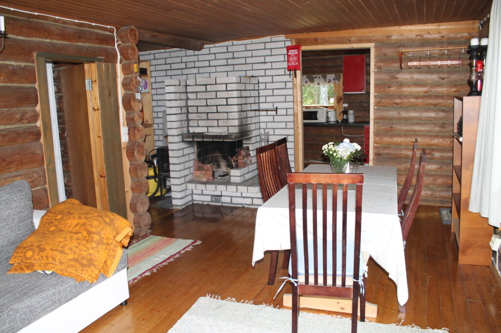 Mökin olohuone perältä päin katsottuna. Kuvassa näkyy ruokapöytä ja takka sekä keittion ovi